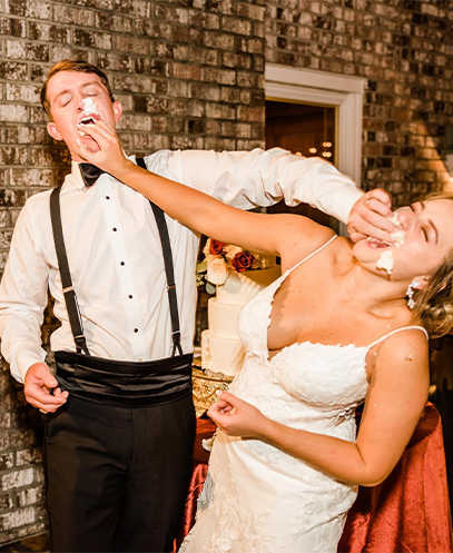 Bride and groom laughing while smashing wedding cake into each other's faces at reception.